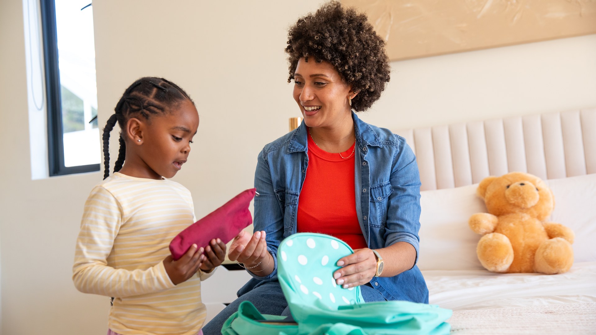 Mother and young daughter packing a backpack with a teddy bear on a bedroom bed before summer camp.