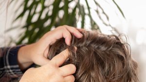 A mother's hands carefully parting a school-aged child's hair near the scalp under good light, checking the part line for nits, dandruff, or other white specks during an at-home identification check.