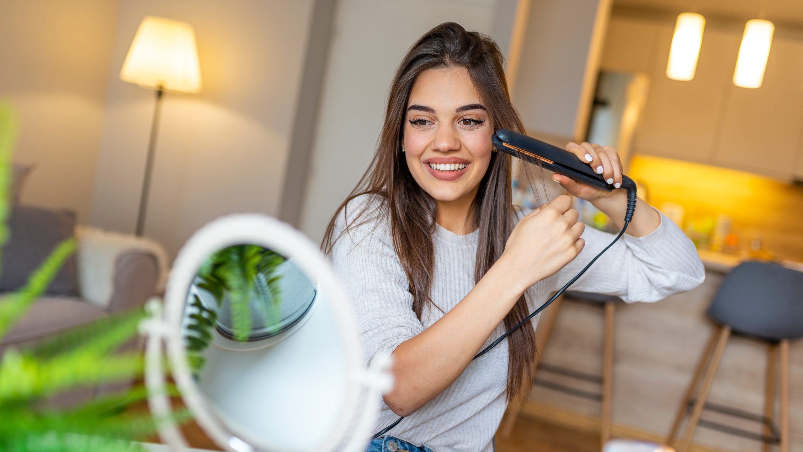 Young woman using a hair straightener flat iron on her hair in front of a bathroom mirror, the visual cue for the moment Broward parents reach for a heat-styling tool to try to kill head lice