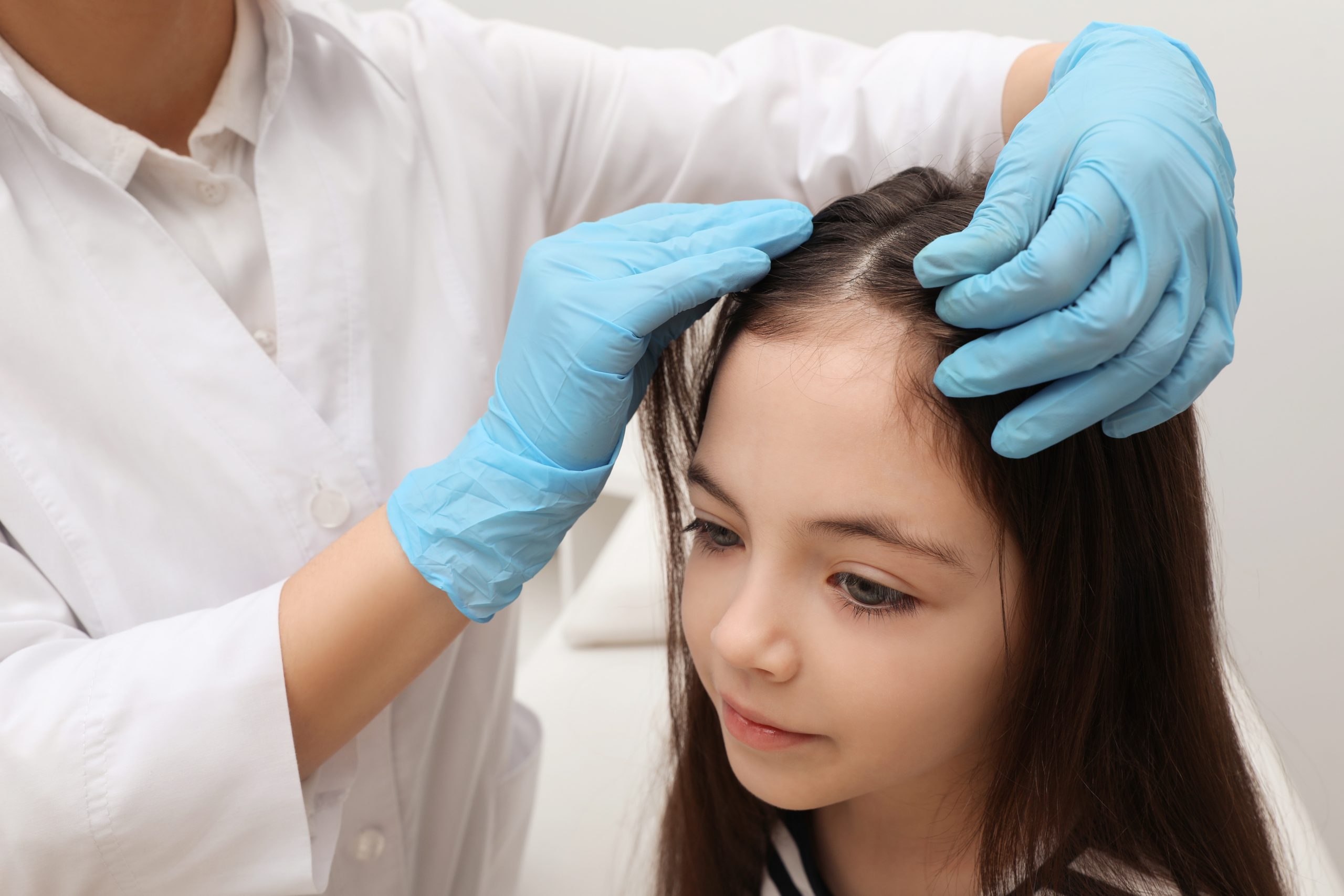 A health professional parts and examines a young girl's hair for head lice under bright light, illustrating the decision of who to see when a child has lice in Broward County
