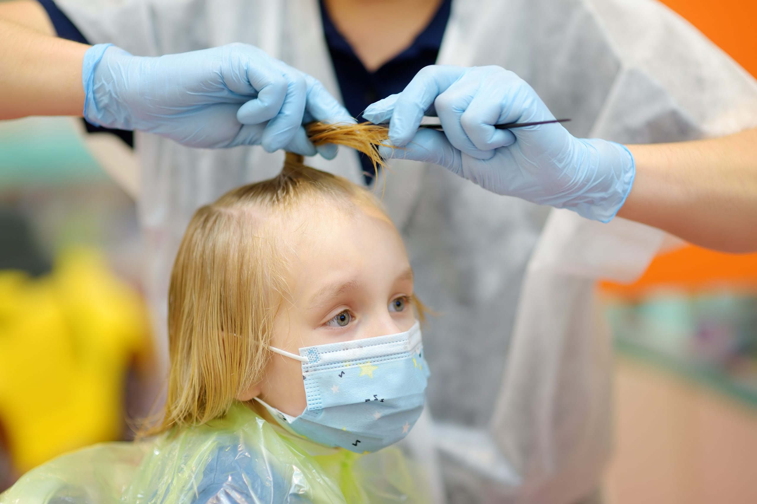 Preschooler boy wearing facemask getting haircut in salon during coronavirus epidemic. Hairdresser examined and treating baby hair for lice.