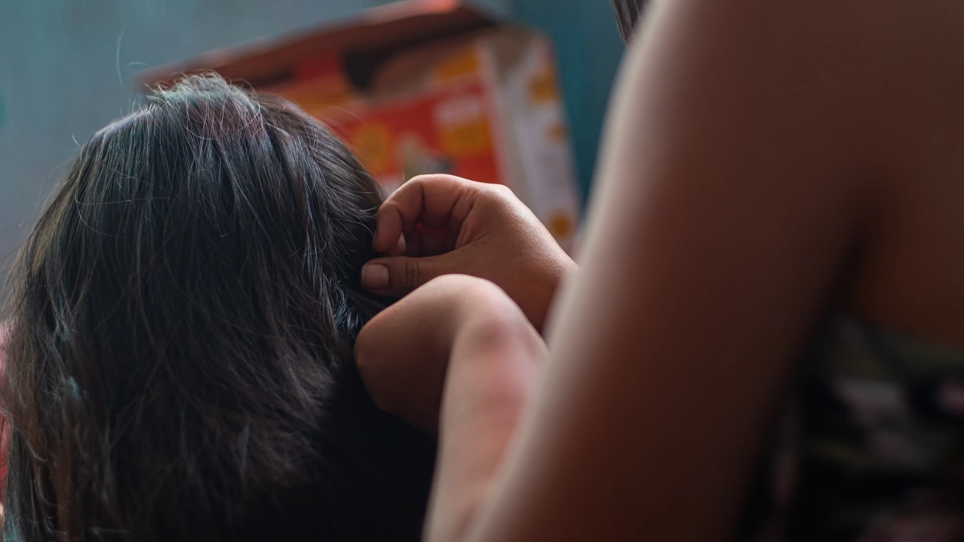 Close-up of a hand lifting a section of long dark hair at the side of a head, with another person's bare arm blurred in the foreground and a colorful box out of focus behind.